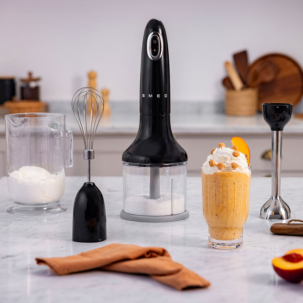 Black Smeg hand blender on a kitchen counter with a glass of dessert and kitchen utensils.