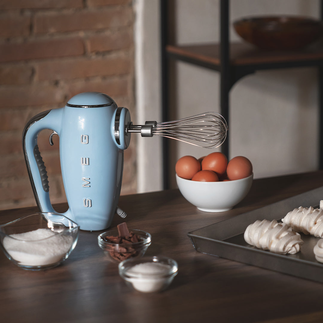 Blue Smeg hand mixer on a kitchen counter with eggs and baking ingredients.